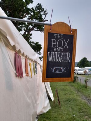 Photo of a wooden sign with chalkboard in the centre, made up to look like a pub sign for a pub called The Box and Whisker, with a box and whisker plot drawn on it. The sign hangs off the side of a white tent with the word 'MATHS' visible made from coloured wooden letters hanging along one side